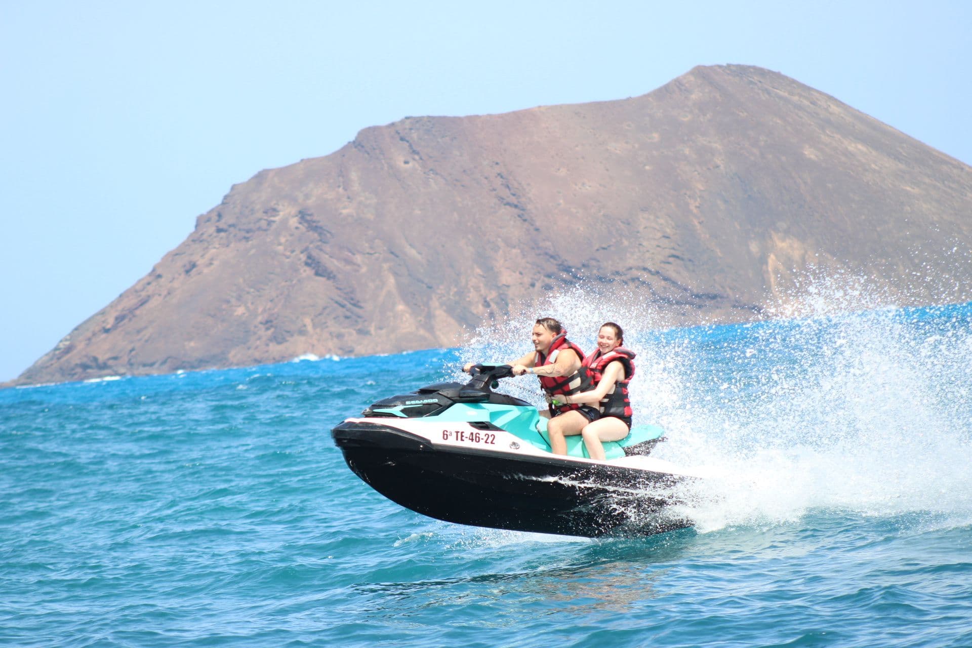 Couple on a turquoise jet ski below the Montaña La Caldera volcano on Isla de Lobos, Fuerteventura